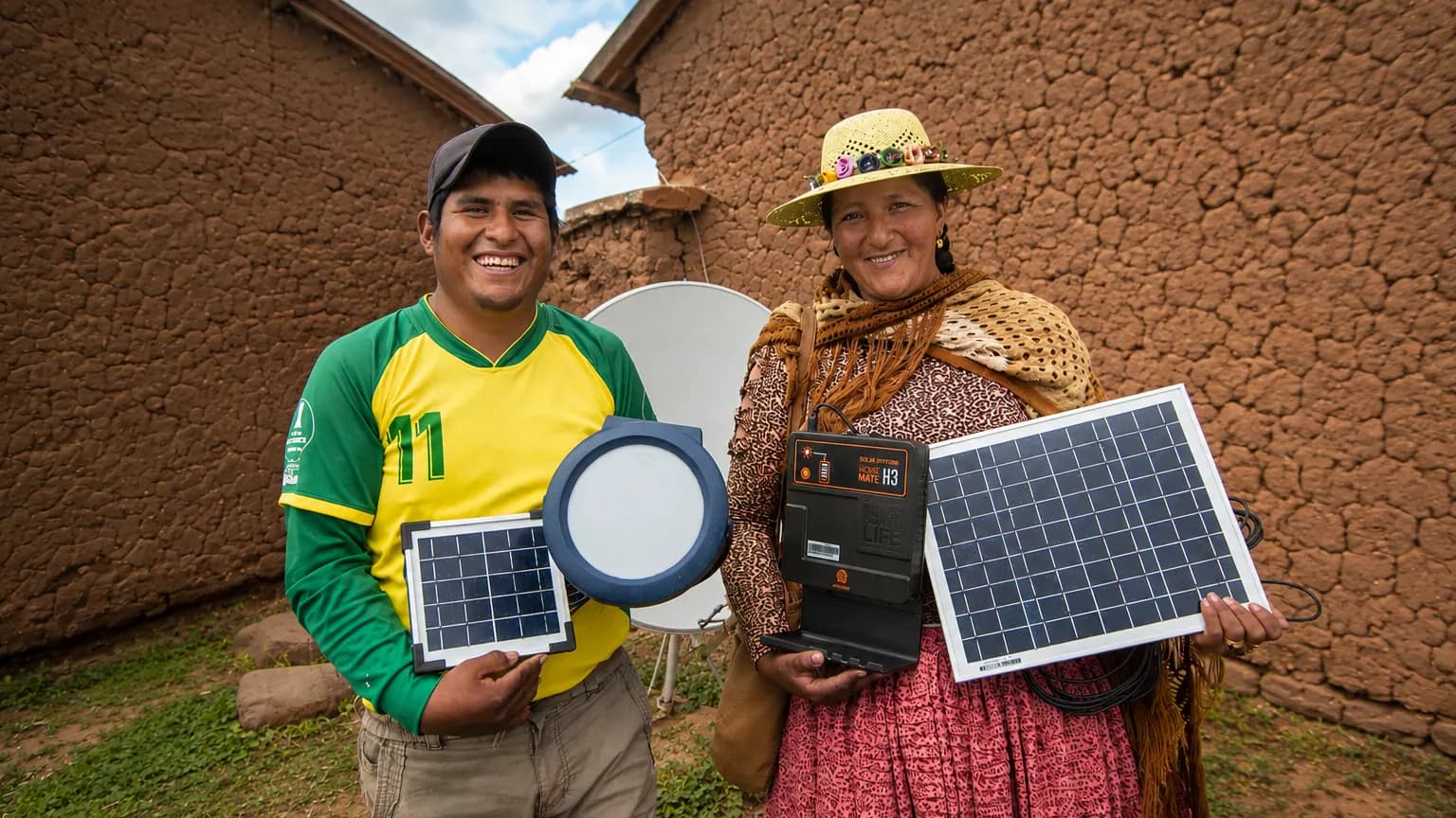 Pareja aymara sosteniendo paneles solares y un equipo de energia limpia frente a su casa