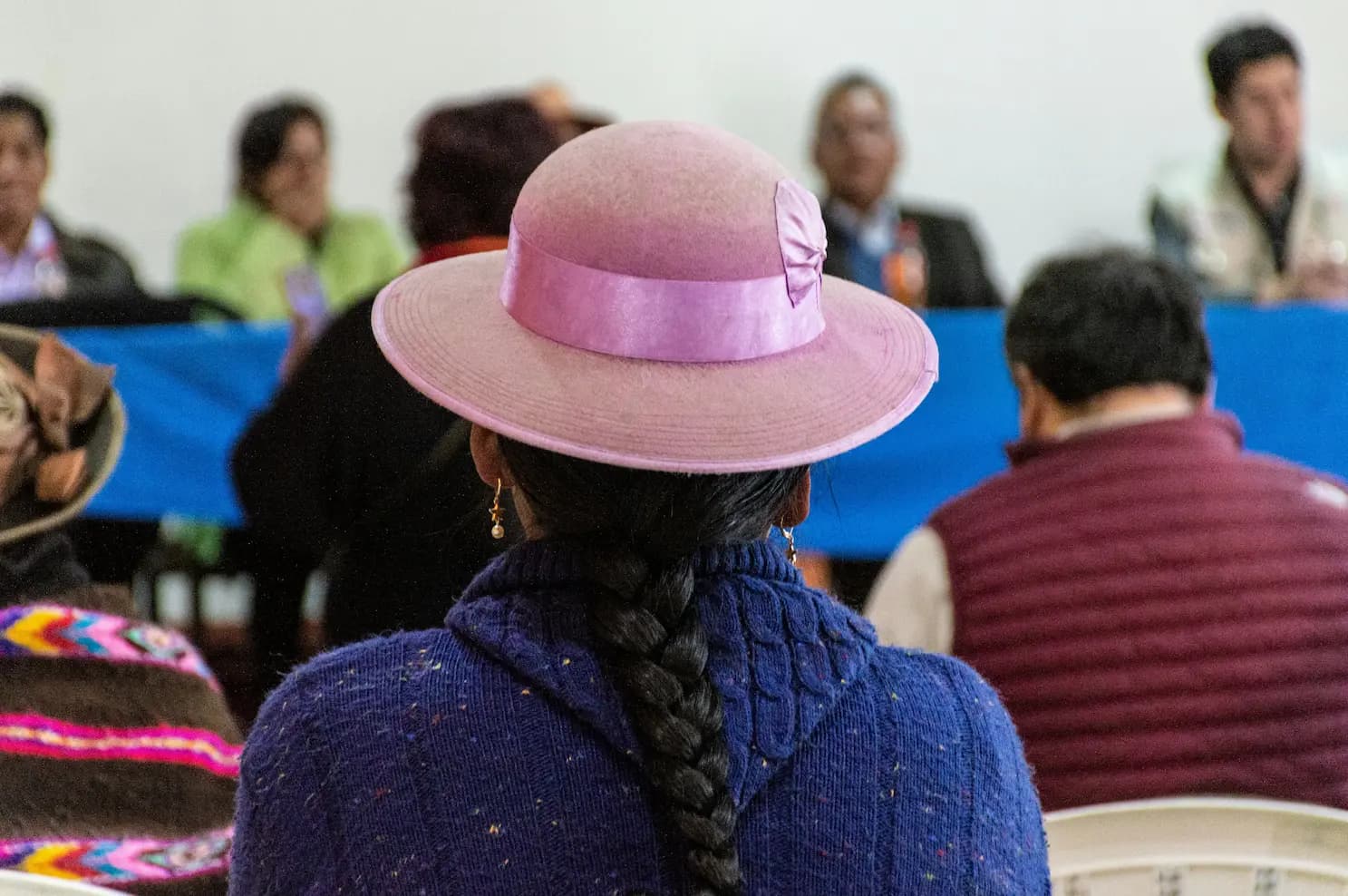 Mujer indígena con sombrero rosa y trenza, de espaldas, en una reunión comunitaria
